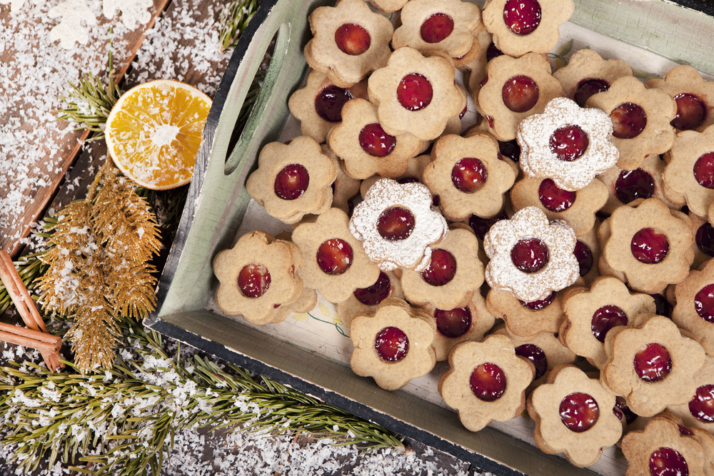 Flatlay of flower shaped vegan Christmas cookies with jam in the middle on a tray next to a fir branches and fake snow.