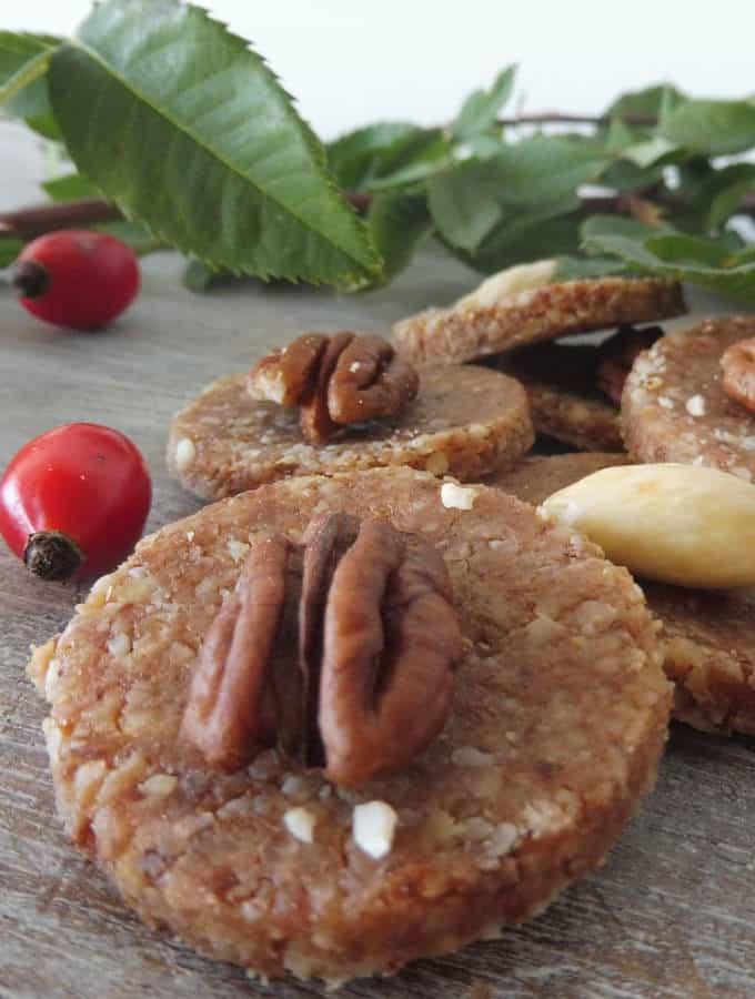 Round, dark brown rose hip cookies with a pecan on top next to leaves and cranberries.