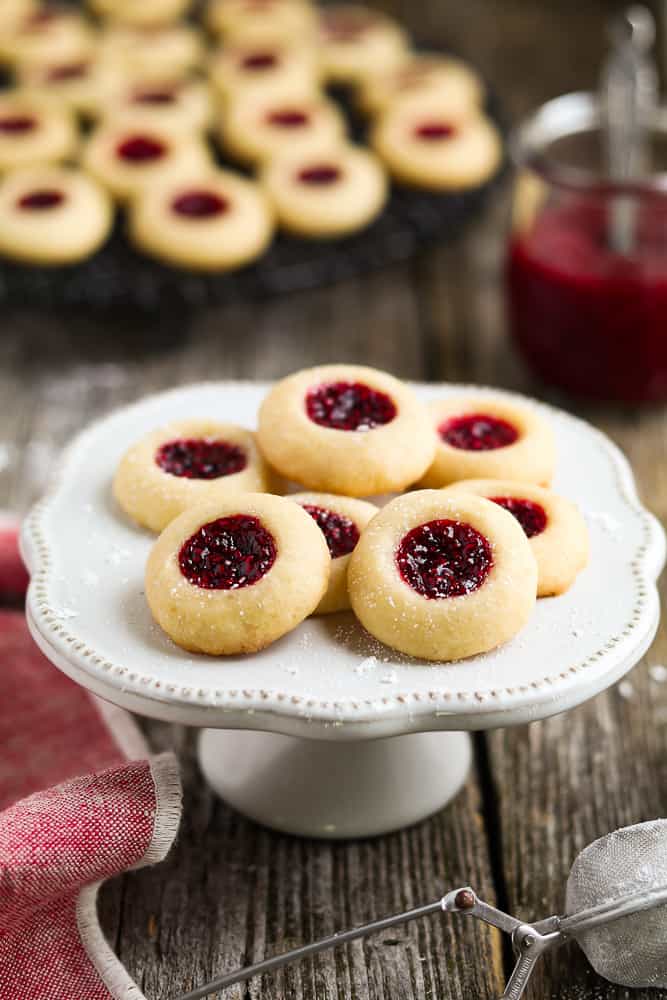 Round, light brown thumbprint cookies with red jam in the middle plied on a raised white tray.