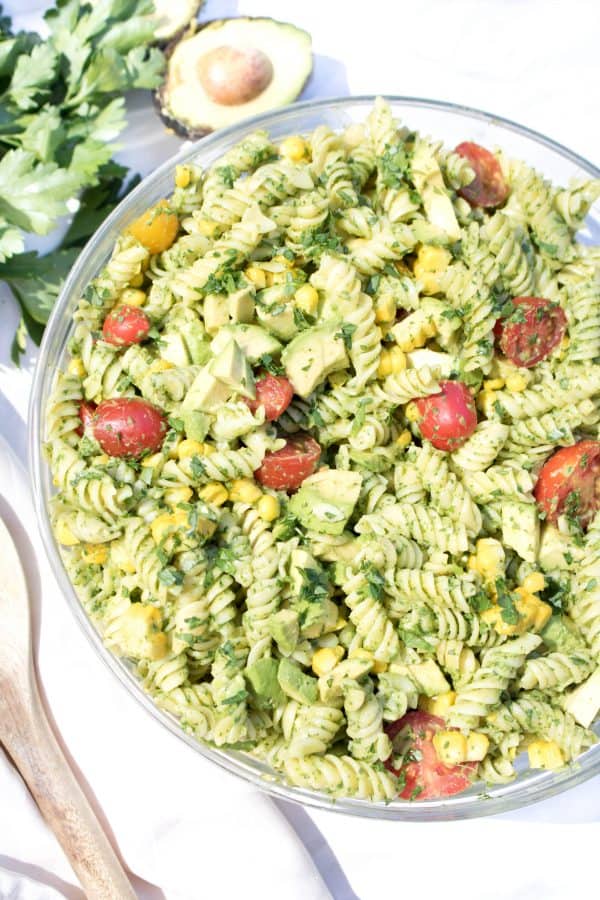 Photo of a large bowl of avocado pasta with a large wooden serving spoon on the left side of the bowl. Cilantro garnish and half of an avocado are seen in the background.