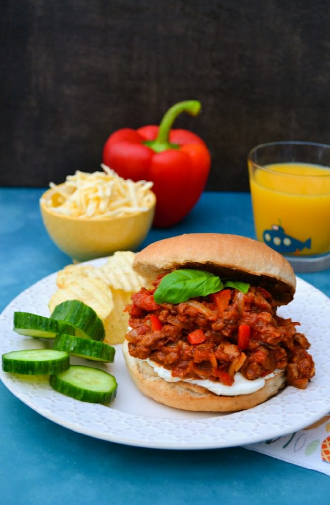 Photo of vegan sloppy joes. A white plate is seen with a vegan sloppy joe on it. Sides of sliced cucumber and potato chips are also on the plate. 