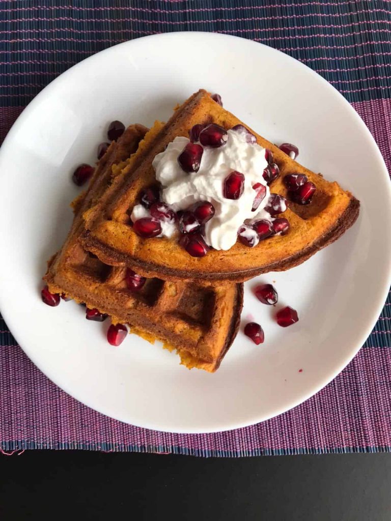 Photo of sweet potato waffles being served with vegan whipped topping and pomegranate pieces on a circular white plate that is placed upon a purple and blue striped place mat. 