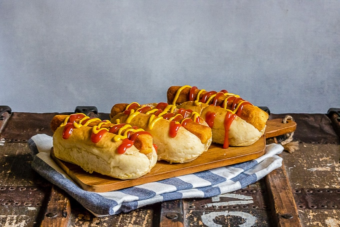 Photo of three vegan hot dogs in buns on a wooden serving board. The board in resting on a blue and white cloth on a rustic trunk.
