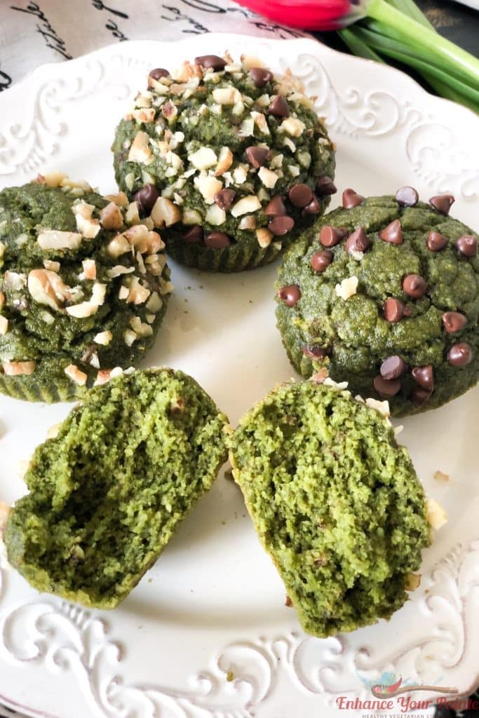 Photo of four vegan banana oats spinach muffins on a white plate. The muffins are an intriguing green color from the spinach and topped with chocolate chips and nuts.