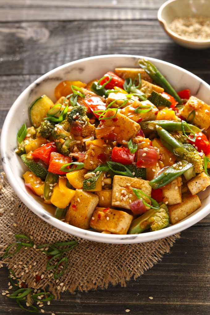 Photo of vegan tofu stir fry. Features a marble bowl with colorful vegetable stir fry with chunks of tofu on a wooden table.  