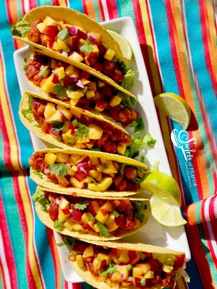 Photo of vegan chickpea tacos with peach salsa being served on a white rectangular plate with lime wedges and a very colorful striped tablecloth.