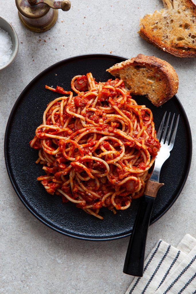 Photo of Vegan Lentil Bolognese on a black dish with a silver fork. A chunk of hearty bread is on the side of the plate. 