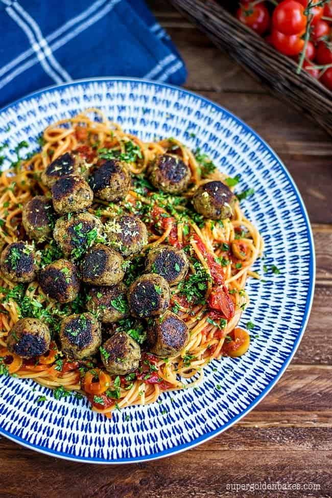 Photo of vegan aubergine "meatballs". A blue and white plate is serving the dish. There is a basket of vine tomatoes and a blue with white stripes cloth napkin in the background.