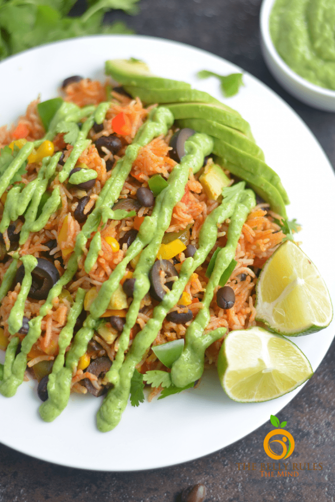 Photo of vegan burrito bowl being served on a white round plate. Lime wedges are on the side of the plate with a bright green sauce drizzled on top.