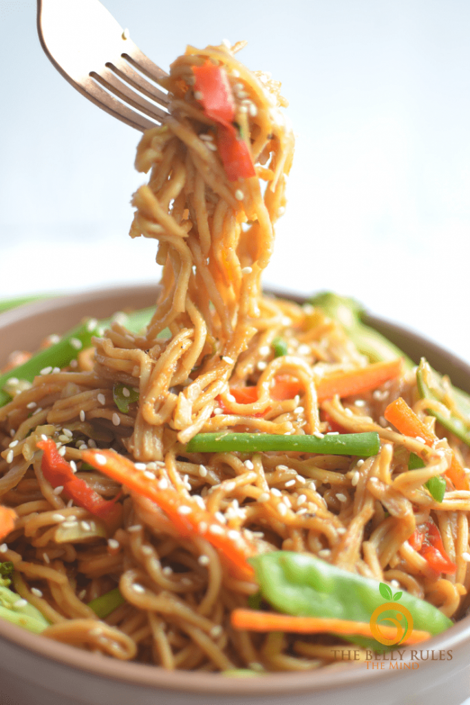 Photo of vegetable chow mein. A fork is seen with noodles dangling into the bowl. Some vegetables are seen as well as sesame seeds. 