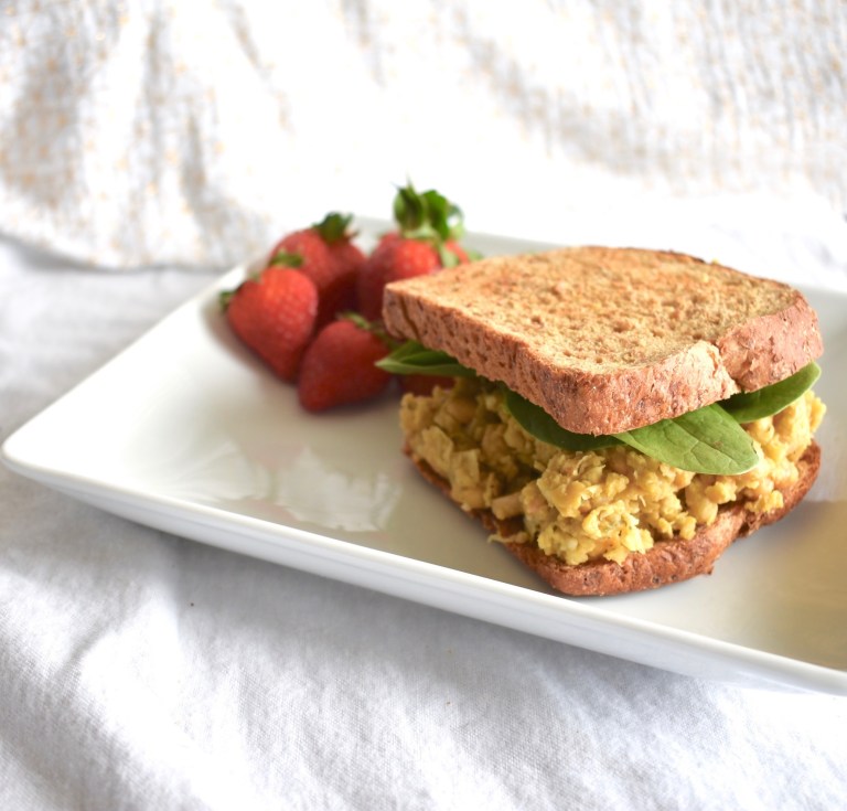 Photo of vegan buffalo chicken salad sandwich being served on a white square plate with a side of fresh strawberries.