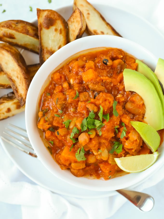 Photo of vegan chilli with butternut squash and chipotle being served in a small round white bowl with avocado slices as a garnish. There is also a side of potato wedges. 
