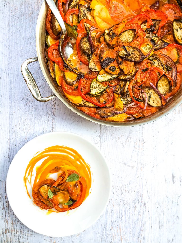 Photo of Vegan French Ratatouille being served from a large steel pot with a large serving spoon. The pot is on a white washed wooden surface.