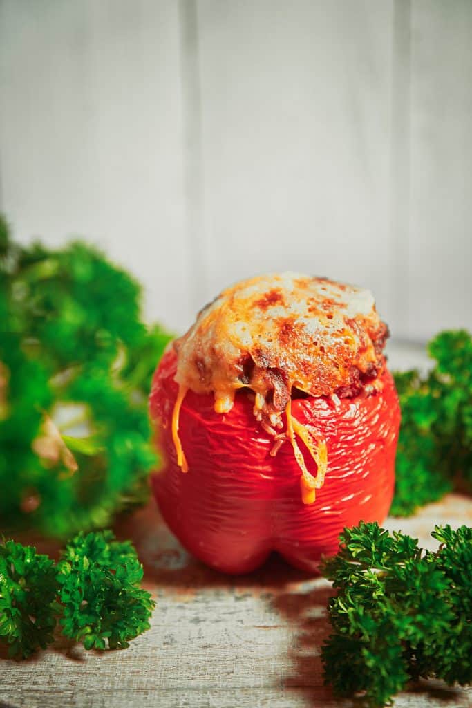 Close up of a single baked, red stuffed bell pepper with melted vegan cheese on top surrounded by parsley.