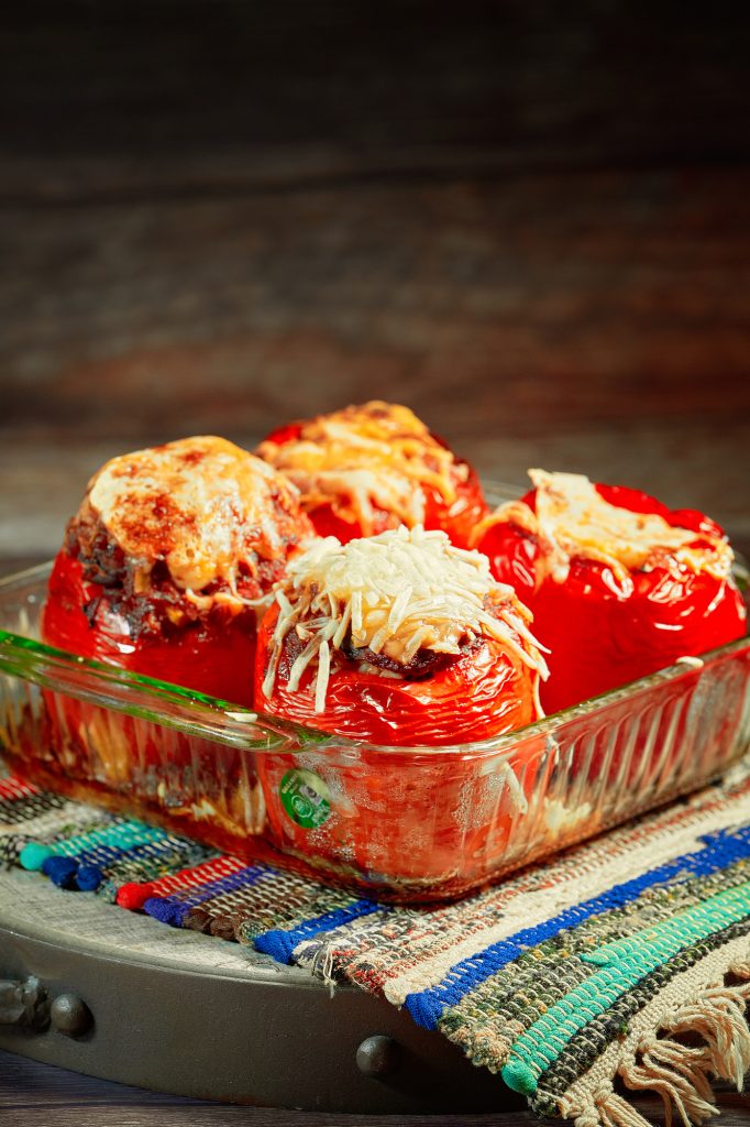 Baked vegan stuffed red bell peppers in glass baking dish colorful mat on wood background.