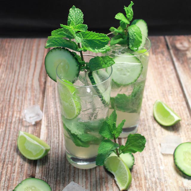 Photo of two light and fresh cucumber mojitos with cucumber slices and mint sprigs as a garnish on a wooden table with more limes scattered around.