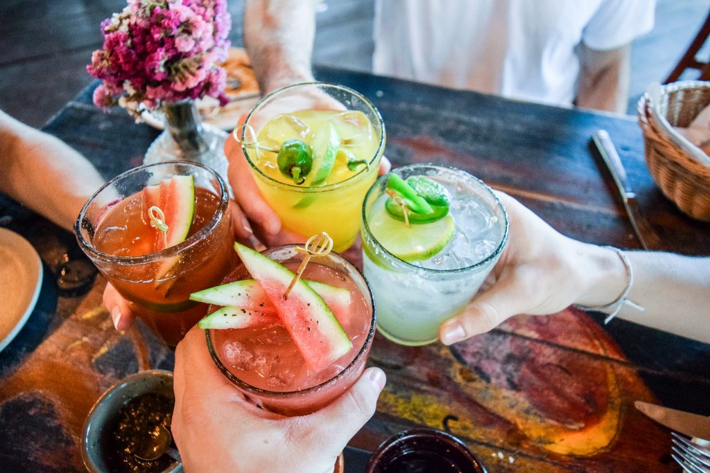 Photo of four different colorful cocktails in glasses being brought together in a cheers clinking motion.
