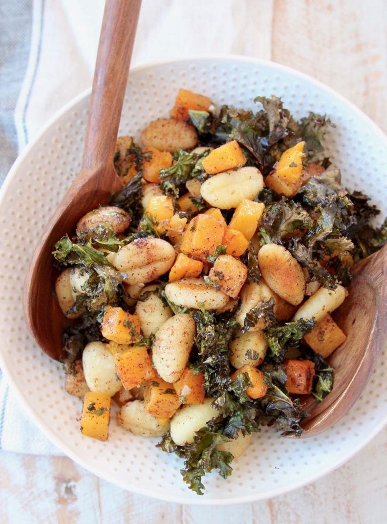 Photo of baked gnocchi with butternut squash and crispy kale being served in a white bowl with a textured rim with a wooden spoon.
