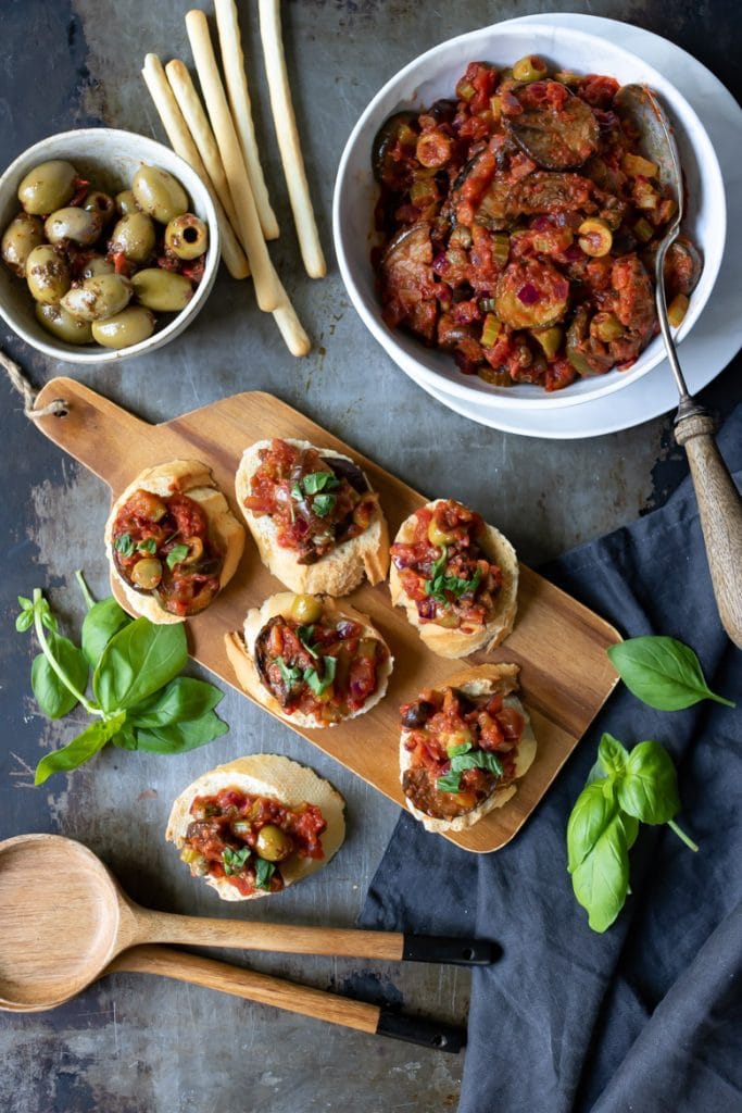Photo of a wooden board serving caponata alla siciliana on crostinis next to a large bowl on the dish and a small bowl of olives.