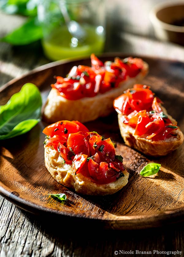 Photo of three baugette slices topped with bruschetta with basil-infused olive oil being served on a round wooden plate.