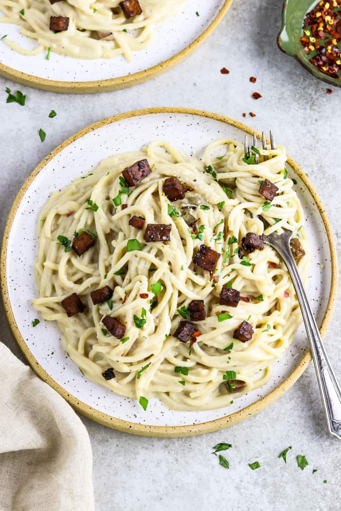 Photo of a rustic plate serving vegan carbonara. A small dish of red pepper flakes is off to the side.