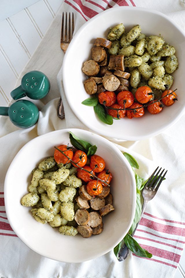 Photo of two white round bowls serving gnocchi with pesto, roasted tomatoes, and vegan sausage. This recipe proves that vegan Italian recipes can look complex and still be easy. 