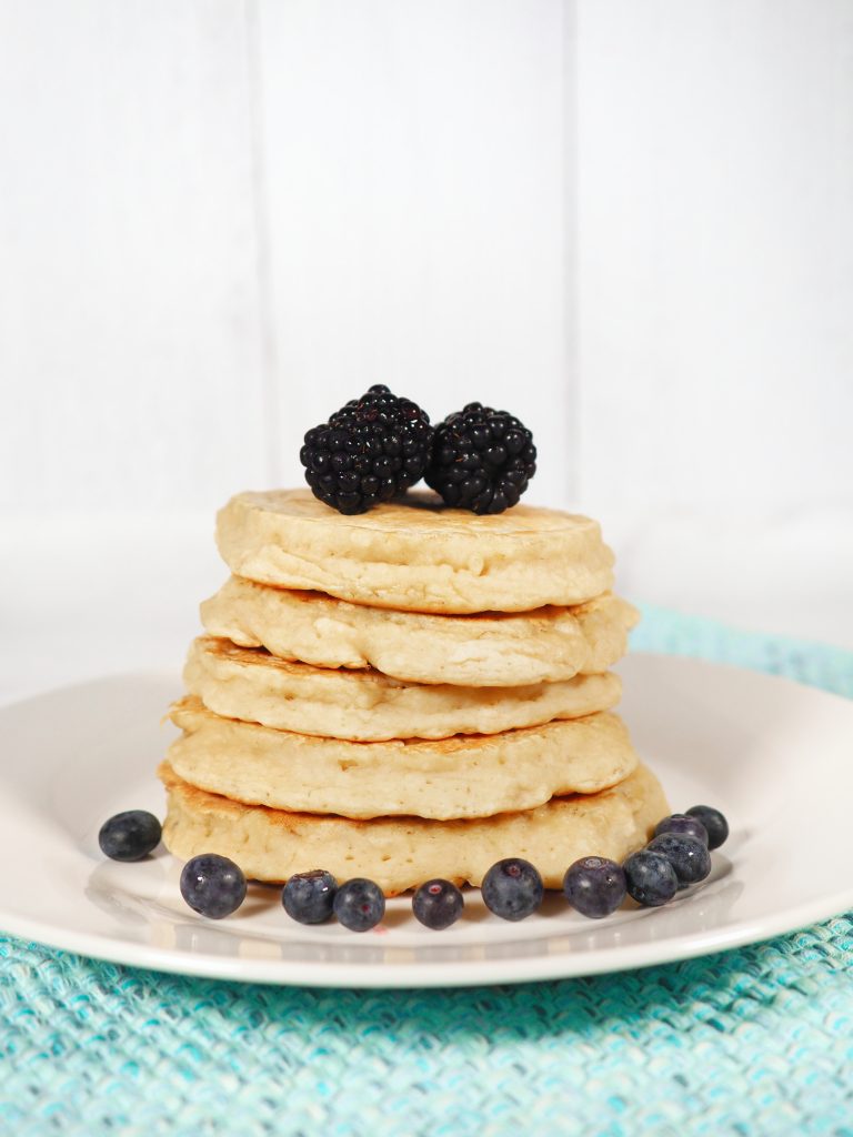 Photo of 5-ingredient vegan banana pancakes being served on a round white plate that is sitting upon an aqua woven place mat.  There are 5 pancakes stacked on top of each other with blueberries and blackberries.