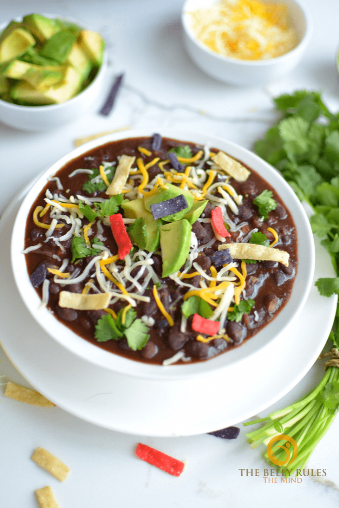 Photo of spicy black bean soup being served in a round white bowl with avocado, cheese, and tortilla strips as a garnish.