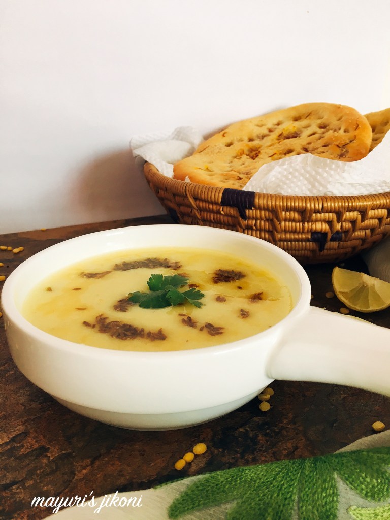 Photo of Moong Dal Shorba/Moong Dal Soup being served in a white bowl with naan bread in the background. One of the more unusual soups among the vegan soup recipes. 