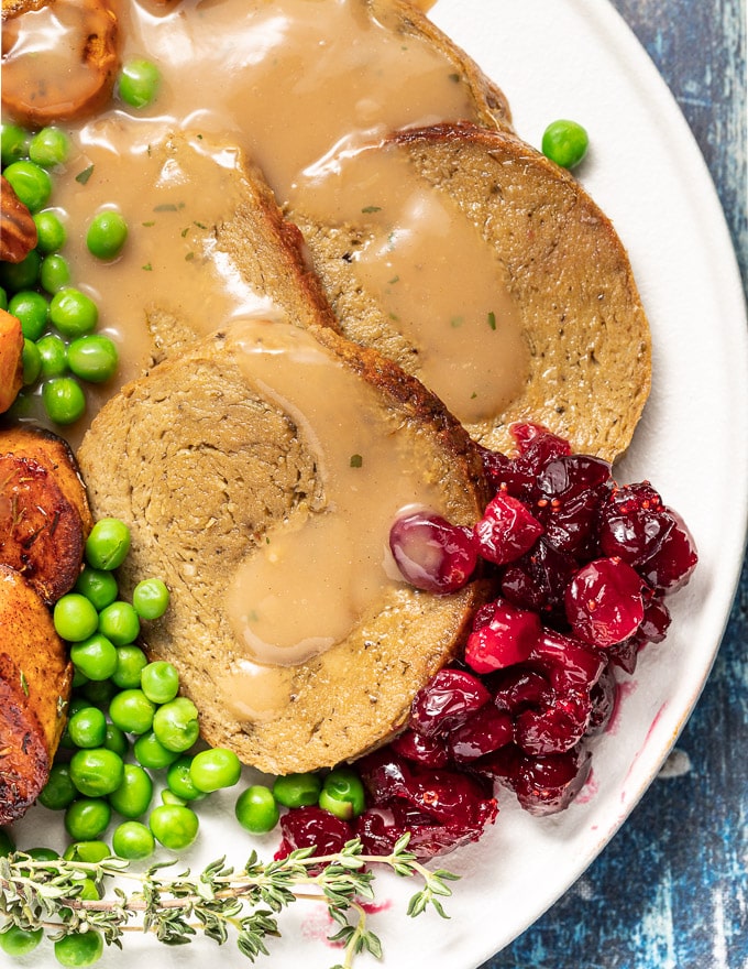 Photo of a sliced vegan roast with gravy on top, also being served with peas and cranberries. 