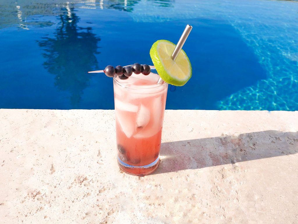 Photo of a blueberry vodka lemon spritzer with a lime slice and blueberry skewer garnish on the edge of the outdoor pool.
