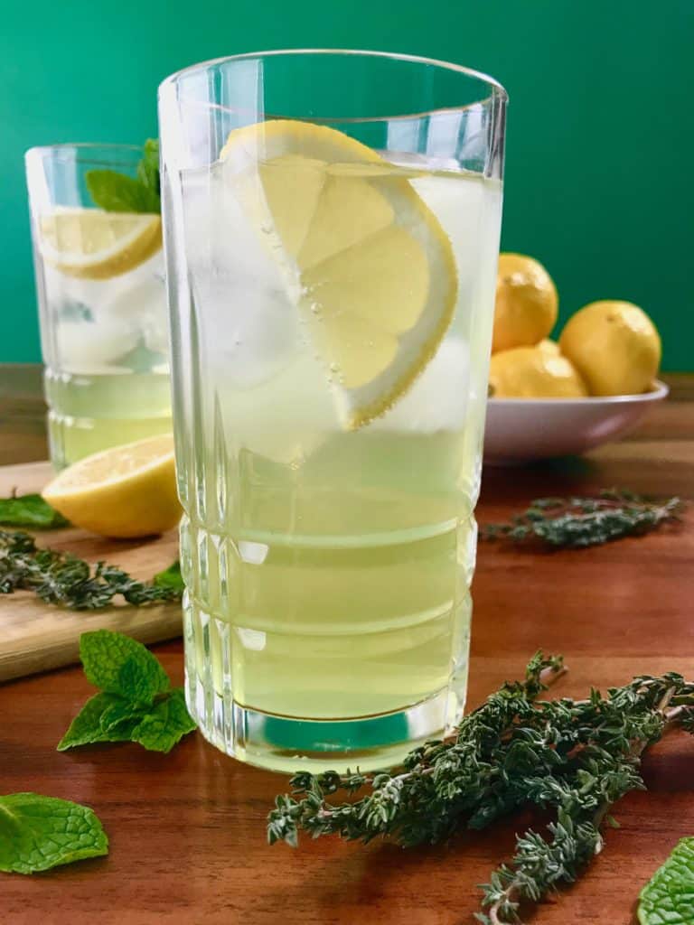 Photo of two tall glasses serving a limoncello spritz with ice and a lemon garnish next to a bowl of lemons and scattered mint.