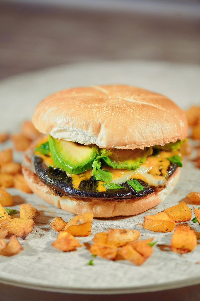 Photo of vegan portobello mushroom burger being served on a rustic wooden board with oven roasted paprika potatoes surrounding the burger.