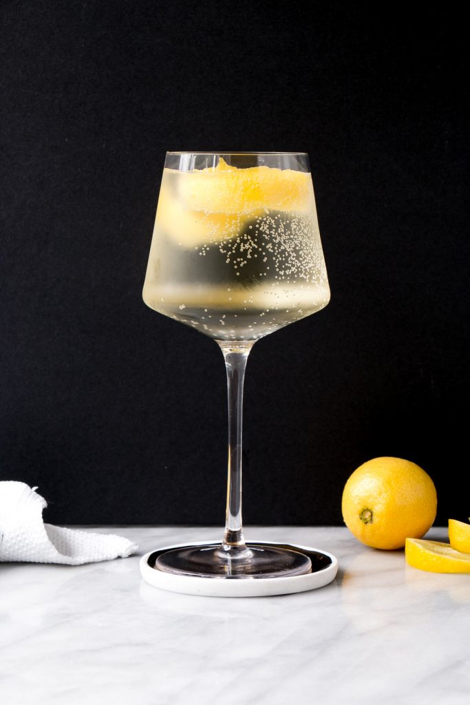 Photo of a white wine spritzer with lemon garnish against a black background on a marble counter next to a lemon.