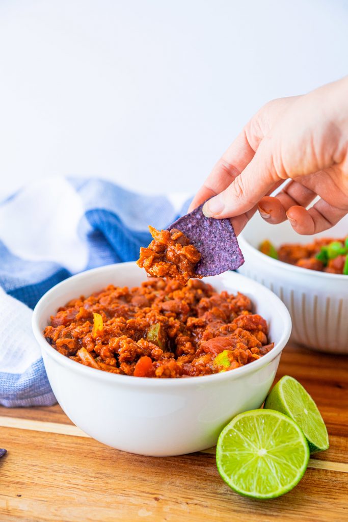 Hand scooping vegan chili with a blue corn chip from a white bowl next to limes.