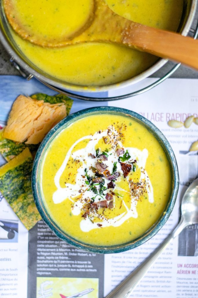yellow color vegan pumpkin soup in bowl with white coconut milk and soup pot with other pieces of orange pumpkin on the table on a newspaper