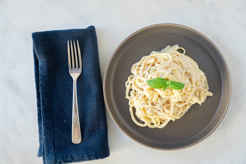 A flatlay image of vegan fettuccine alfredo pasta with fork and a blue napkin.