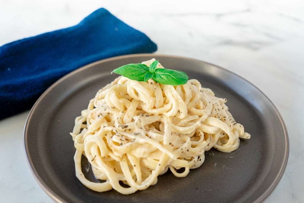 A close up photo of vegan alfredo sauce on pasta on plate with basil leaf next to a blue napkin.