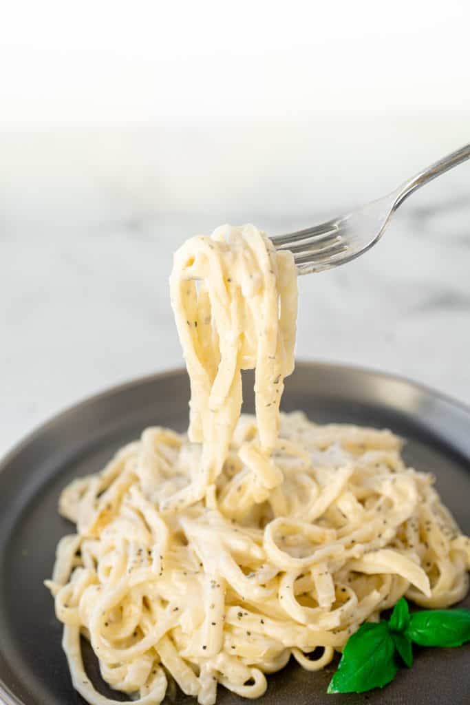 Fork lifting a bite of pasta with vegan alfredo sauce over a plate with basil.