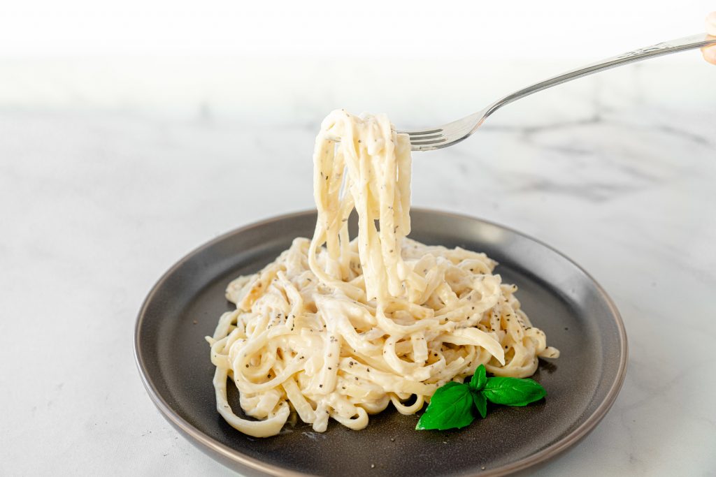 A fork picking up vegan alfredo pasta with creamy sauce from a grey plate on marble counter.