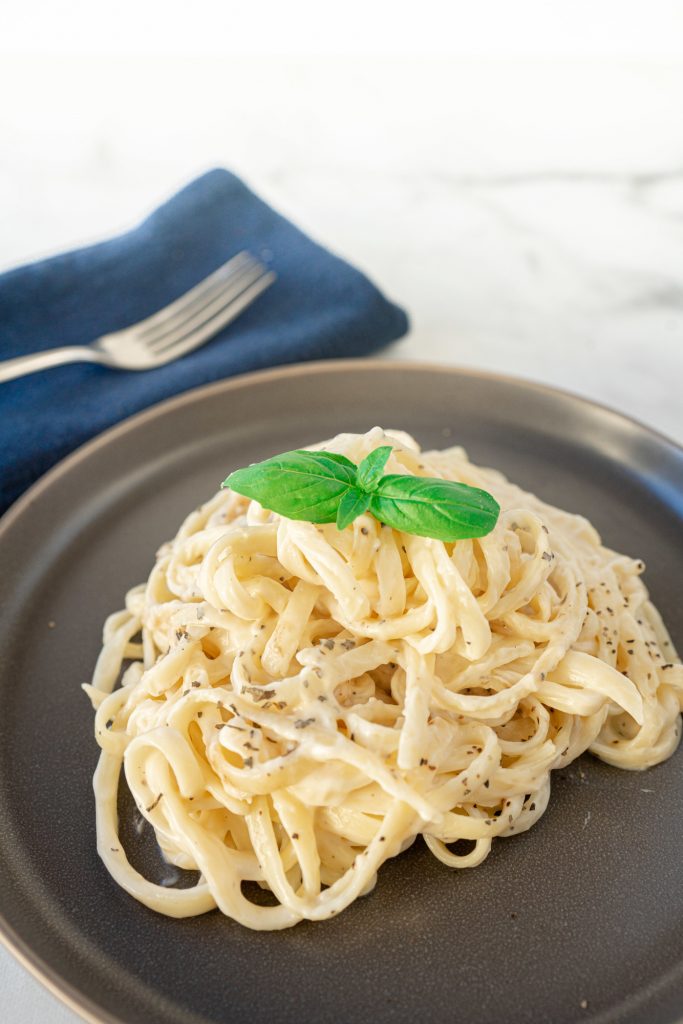 Pile of vegan fettuccine alfredo on grey  plate next to a napkin with fork.