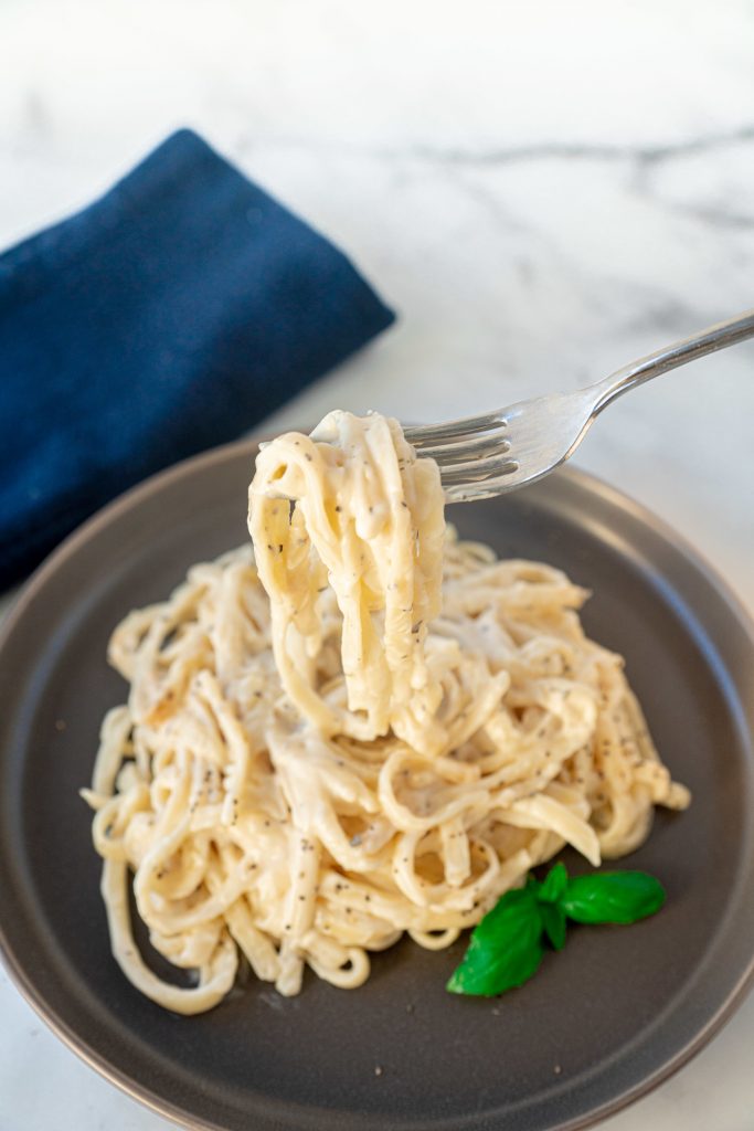 A fork twirling pasta topped with vegan alfredo sauce with no cashews on grey plate.