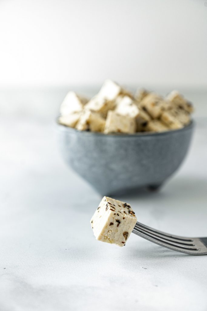 fork holding a slice of tofu feta lying on counter with bowl of feta in the background