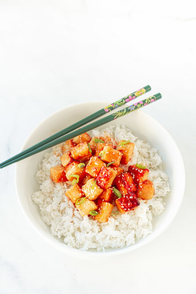 chopsticks resting on the side of a bowl of orange tofu with rice