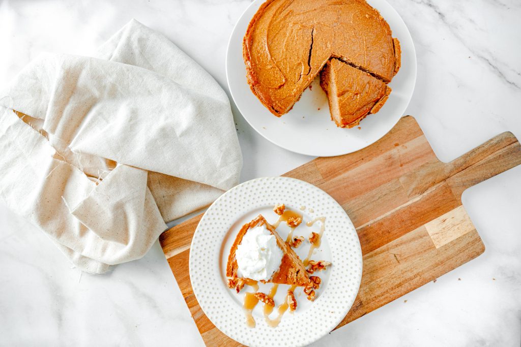 slice of non-dairy pumpkin cheesecake on plate on top of brown bread board with cheesecake in the background