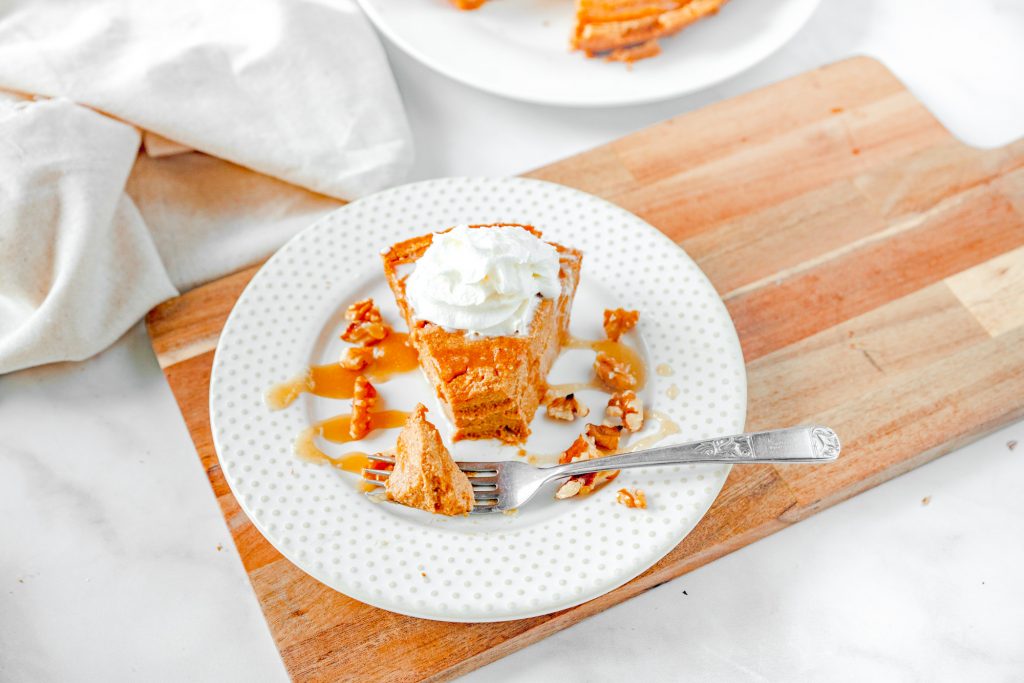 fork with piece of vegan pumpkin cheesecake on it sitting on a white plate on top of a brown bread board on the counter