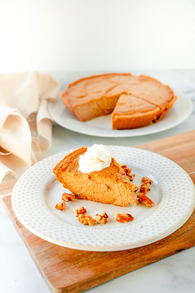 orange non-dairy cheesecake slice on a white plate with full cheesecake in the background with nuts around it