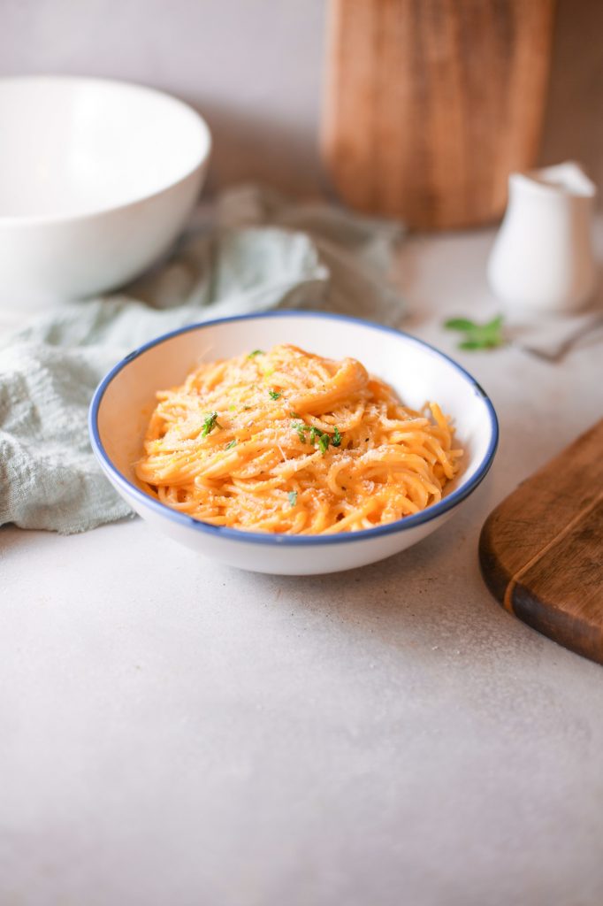 vegan pumpkin pasta sauce over spaghetti noodles in a white bowl with a blue rim and brown breadboard nearby