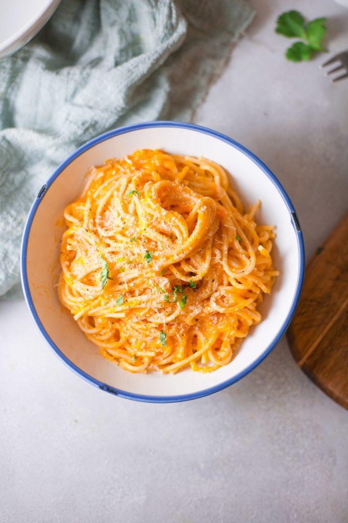 vegan pumpkin pasta in a white bowl on counter with a cutting board. the pumpkin pasta sauce is orange