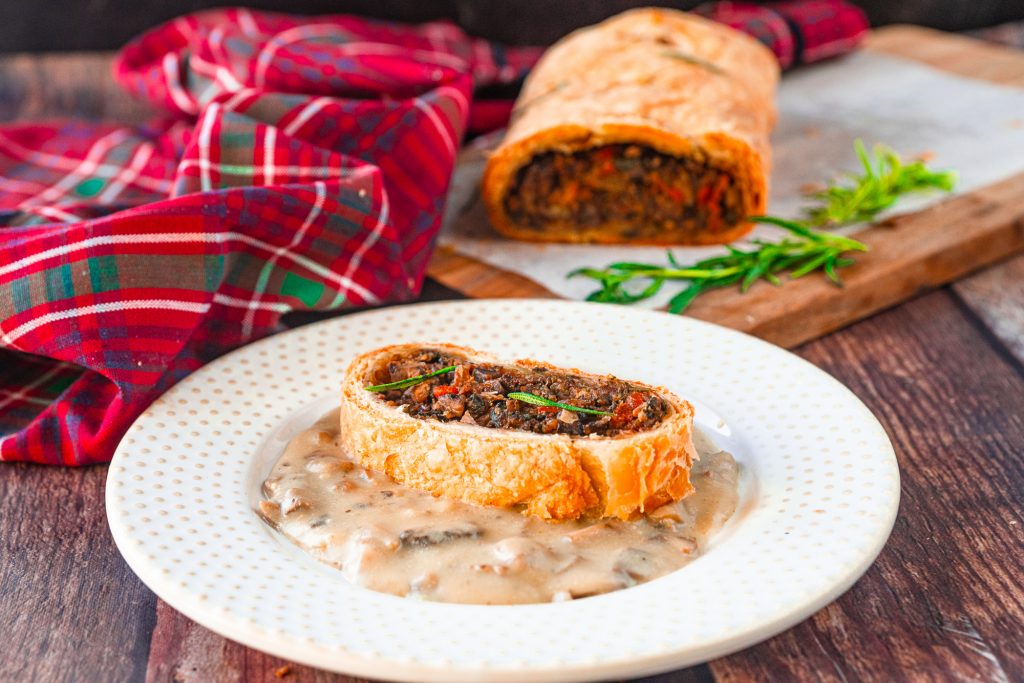 close up of a slice of vegan wellington on mushroom gravy on a white plate with red towel in the background